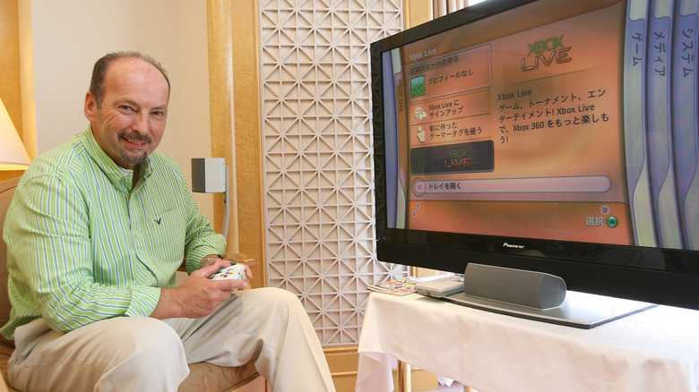 Xbox executive Peter Moore holding a controller in front of an Xbox 360 main menu on TV